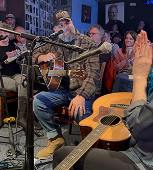 Chris Clayton playing an acoustic guitar, singing during a live performance with audience nearby
