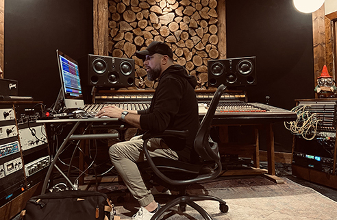 Chris Clayton working at a mixing console and computer in a recording studio with a wooden backdrop