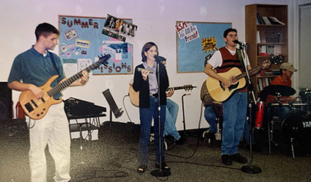 Small band performing indoors, with a singer at a microphone, guitarist, bassist, and drummer