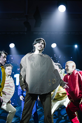 college students in a brown costume standing on stage under spotlights surrounded by other students