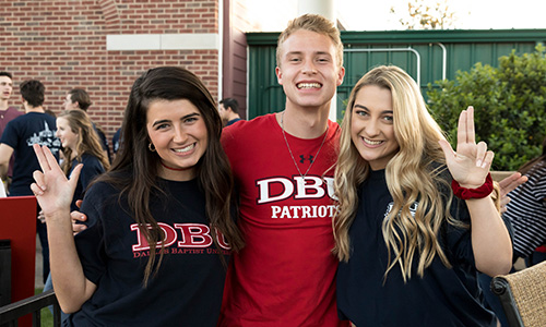 3 students at a baseball game looking at the camera, 2 are making the patriot sign with their hands