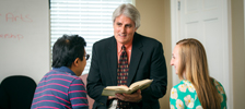 professor talking to two students in classroom while holding a textbook