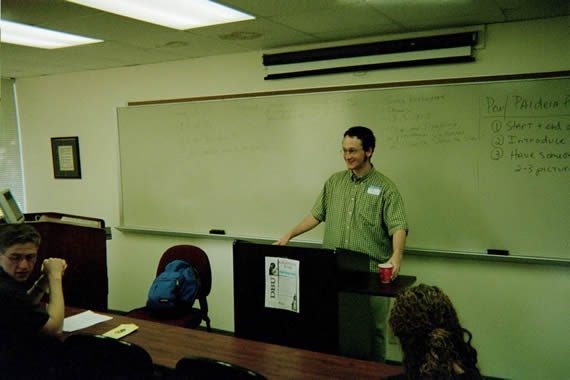 man in glasses wearing a green checkered shirt standing in front of a classroom behind a podium