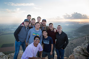 A group of guys posing at the camera on top of a mountain, smiling; The background has clouds and green fields;