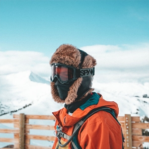 A guy with a black and brown ski mask on, wearing an orange jacket. He’s outside in front of snowy mountains and a wooden fence