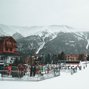 2. A ski resort with snowy mountains in the back. There’s a red and white two-story building to the left and two smaller buildings to the right. In the foreground, there are a bunch of skis sitting outside.
