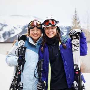 2. Two girls wearing ski gear, posing for the camera together. The girl on the right is blond, wearing a light blue ski jacket. The girl on the right is brunette, wearing a purple-blue jacket.