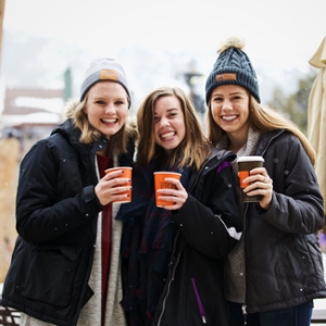 3. Three girls posing tougher outside. The two girls on the right have orange ups in their hands. The girl on the far right and the girl on the far left are wearing snow hats.