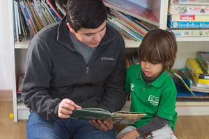 A guy and a little boy reading a book on the floor. The guy is wearing gray Colombia jacket and the little boy is wearing a green collared shirt