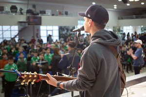 A guy playing the guitar and singing on stage to a group of kids, he’s wearing a hat and a gray sweatshirt