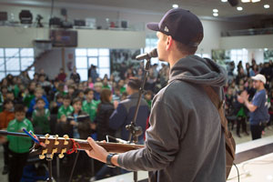 A guy playing the guitar and singing on stage to a group of kids, he’s wearing a hat and a gray sweatshirt