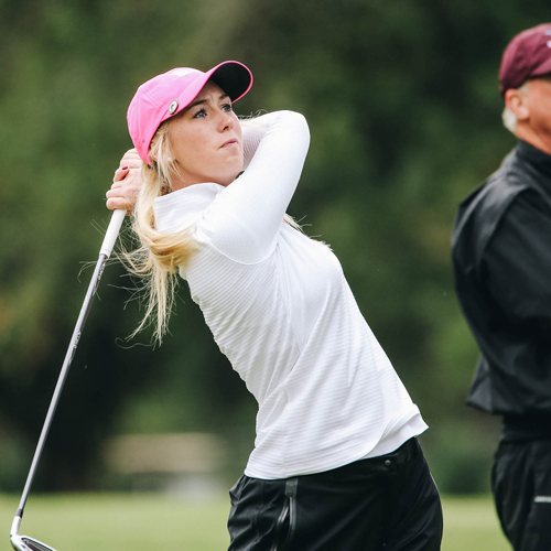 A girl golfer with a white, long-sleeved shirt and a bright pink hat