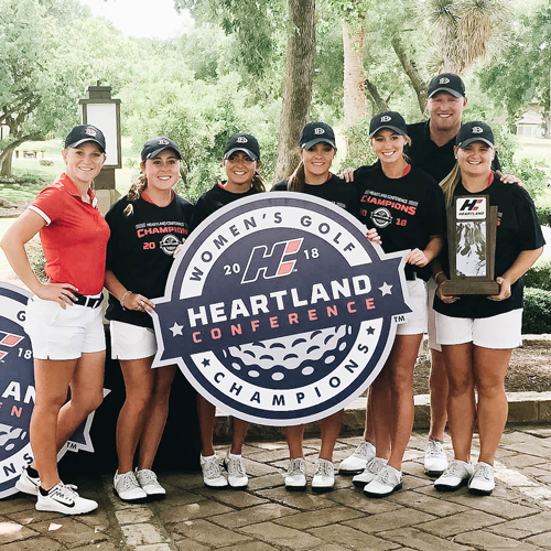 A group of DBU's female athletes posing in front of the Women’s Gold Heartland Conference sign (the sign is circular and blue, and the team is outside)