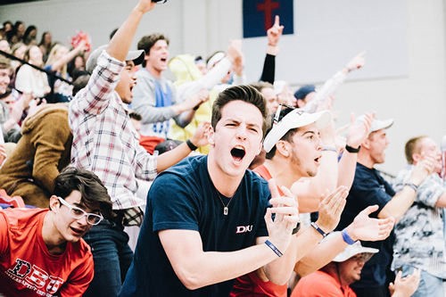 A group of guys cheering in an audience in the basketball gym. The guy in the front is wearing a blue DBU shirt and the guy in the left corner is wearing a red shirt with white glasses at a DBU basketball game