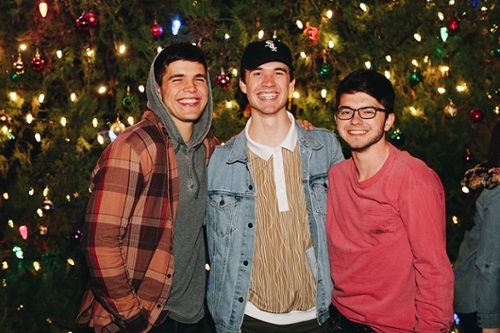 Three guys posing in front of a Christmas tree with lights on it, the guy on the far left is wearing a plaid shirt.
