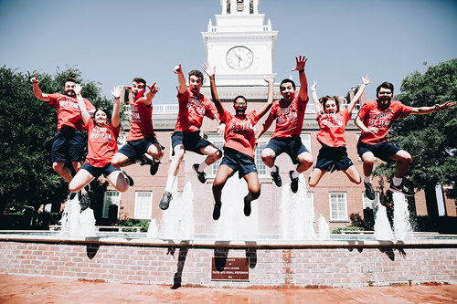 A group of students jumping in the air in front of the fountain outside the Mahler Student Center, all wearing red