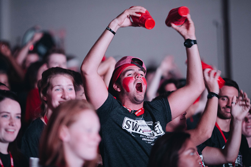 A guy wearing a black shirt in a crowd holding two red cups in the air, he has red face paint on