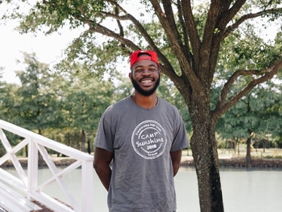 Derrick smiles as he poses in front of Bush Pond with a camp sunshine t-shirt on