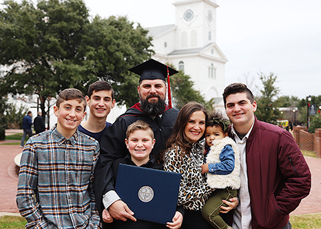 Chris Clayton outside with his family wearing a graduate cap and gown holding his master's degree