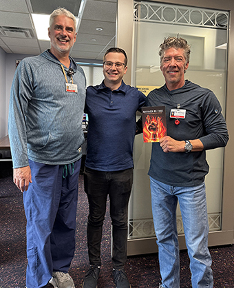 Christian Englert stands indoors with two men; the man on the right is holding up his book.