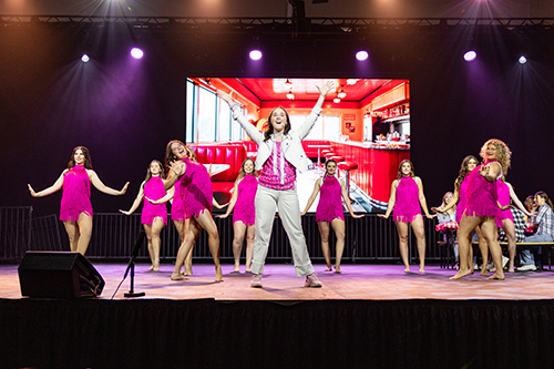 college students singing on stage with dancers wearing pink with a diner scene on the backdrop