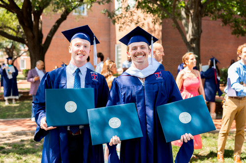 Two graduates in DBU caps and gowns hold diploma folders