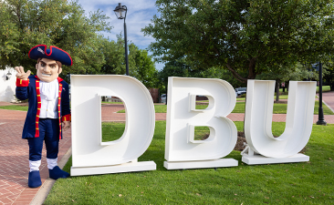 patriot man mascot standing outside by the letters D B U
